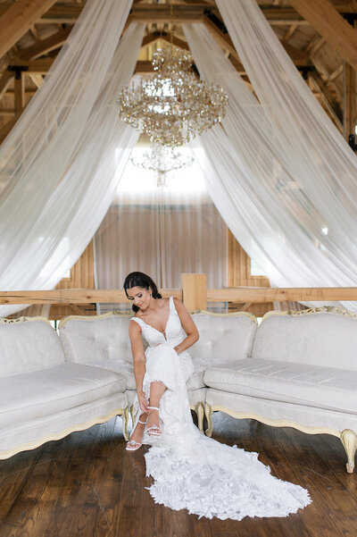 
Bride in a lace gown adjusting her heels beneath a chandelier and flowing white drapes in the loft of a rustic barn at Bear Ridge Destination.

