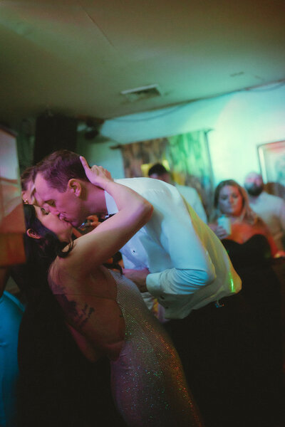 A newlywed couple shares a passionate kiss on the dance floor, surrounded by friends and glowing party lights—captured by an East Coast film wedding photographer.