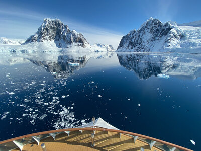 View from the bow of a boat sailing through calm, icy waters surrounded by snow-covered mountains reflecting in the water under a clear blue sky.