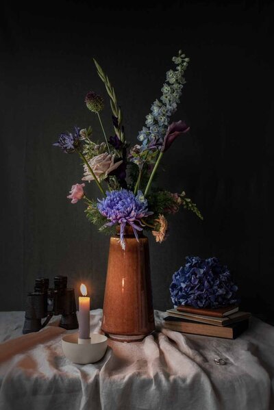 Bloemstuk met blauw, roze en paars gekleurde bloemen in een hoge okerkleurige vaas, met rondom een verrekijker, kaars, trouwringen en notitieboeken