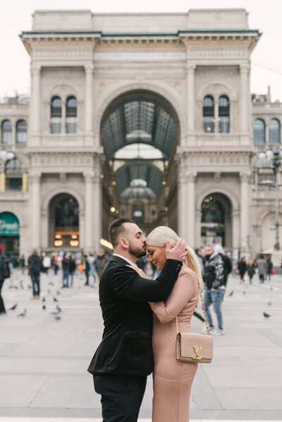 This beautiful photo captures a couple standing in front of the iconic Duomo di Milano in Italy, in a moment of pure joy and love. The man has just proposed, and the woman is overwhelmed with happiness, as he tenderly kisses her on the head. The stunning architecture of the cathedral serves as a stunning backdrop for this romantic moment, which captures the essence of love and the magic of Italy in one unforgettable image