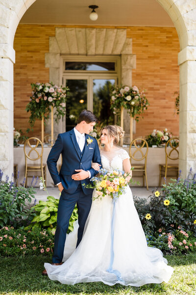 A bride and groom link arms and gaze at each other in front of an elegant center table with large floral centerpieces