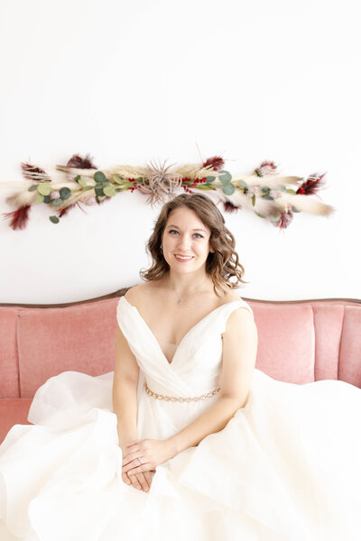 Smiling bride sits on a velvet pink settee in front of a white wall