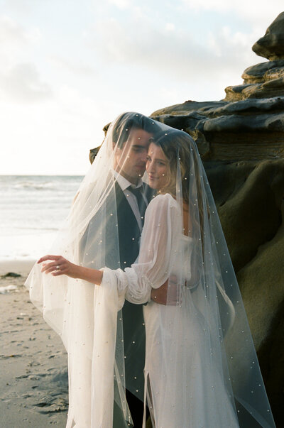 Couple stands under bride's pearl veil in front of large rocks on a beach in San Diego during their elopement.