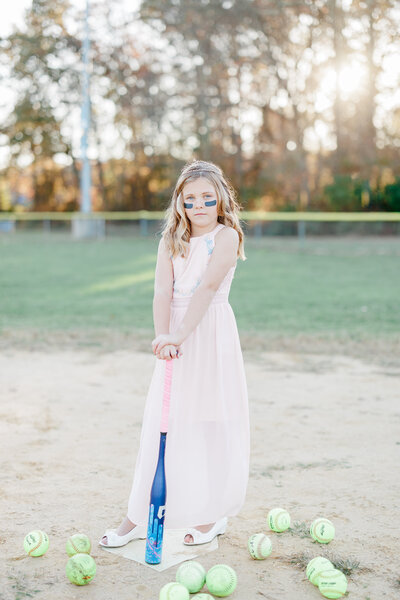 A young girl in a pink dress and white shoes stands on home plate leaning on a softball bat