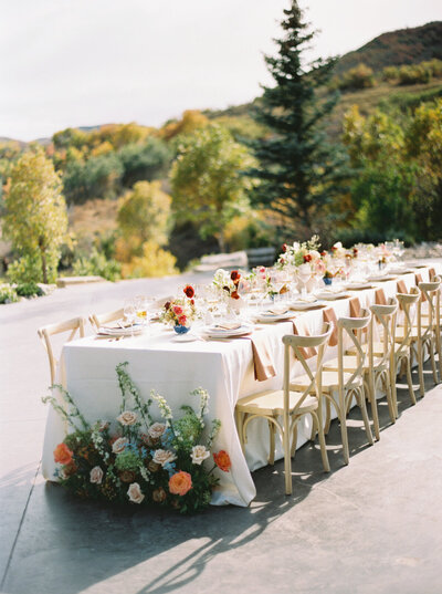 A wedding reception table setup decorated with red, orange, white and blue flowers. Set among the trees with changing autumn leaves.