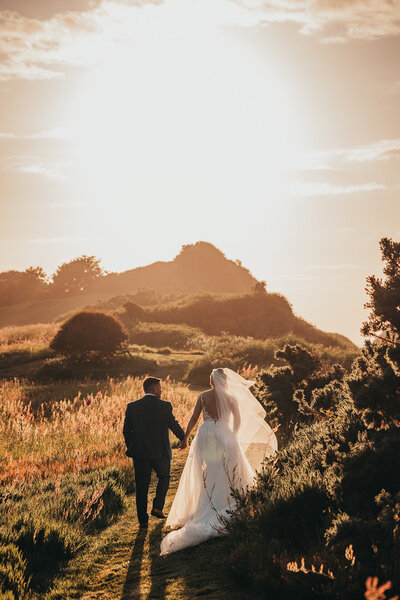 bride and groom hold hands and walk towards sunset