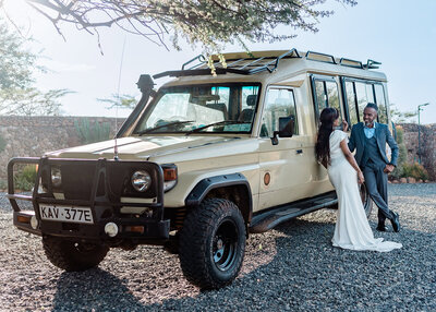 A photographer for luxury elopements in Kenya captures bride and groom in their formal wedding clothes leaning agains a tan colored safari truck talking and smiling. 