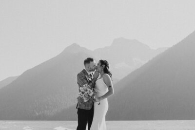 A black and white photo of a couple kissing on a beach surrounded by tall mountains