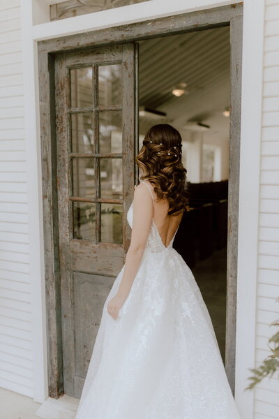 Bride in a white dress with an open back stands by a rustic door, her natural glam wedding makeup enhancing her serene expression as she gazes into a softly lit room.
