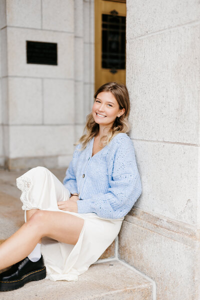 Senior girl in a blue sweater and white skirt sitting and leaning against a concrete wall