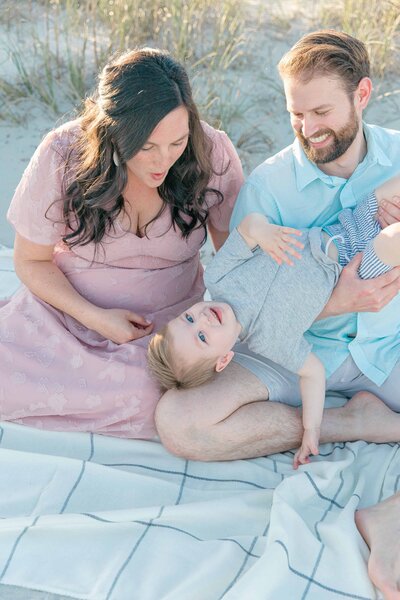 A small family sitting on a beach blanket tickling their toddler wearing light colored clothes for the beach