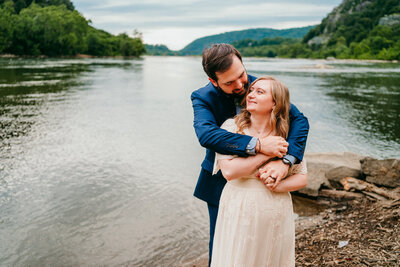 A Couple hugging by the river during their laid-back West Virginia elopement surrounded by green hills.