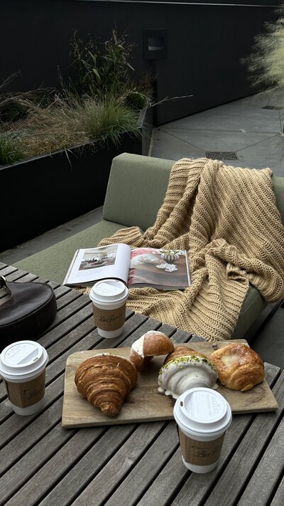 Cozy outdoor scene with a knit blanket on a green sofa, an open magazine, three coffee cups, and pastries including a croissant, on a wooden table.
