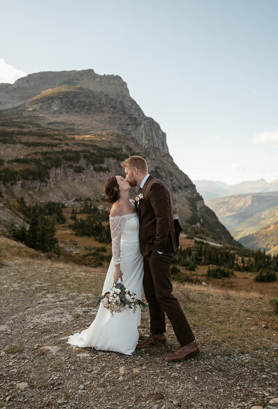 A bride and groom share a kiss on a mountain path surrounded by rugged peaks and soft evening light during their Glacier National Park elopement, captured by Sydney Breann Photography.