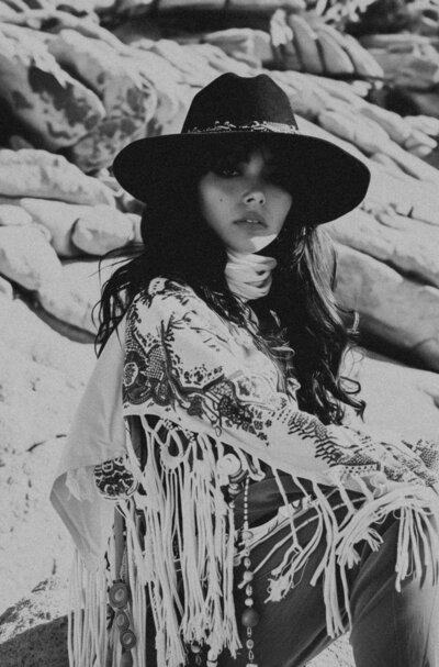 A woman in a wide-brimmed hat and patterned shawl sits on rocks, looking at the camera with a sense of brand identity. The black and white image captures her essence with artistic flair, akin to Sweet Pea Ink Creative's signature style.