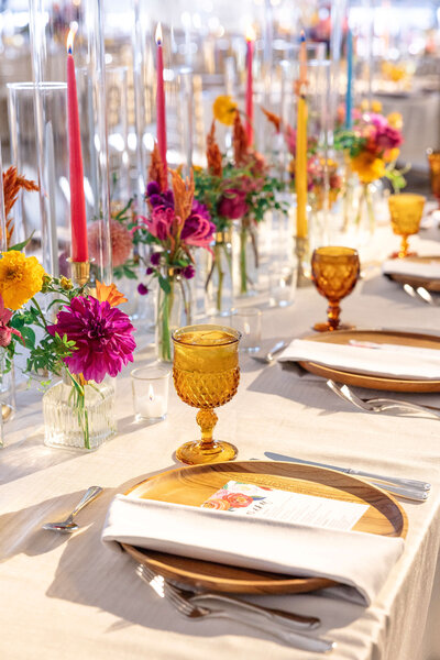 A beautifully set table with wooden plates, amber glasses, and colorful floral centerpieces. Tall red, pink, and yellow candles are arranged in glass holders, adding a touch of elegance. White napkins wrap cutlery on each plate.