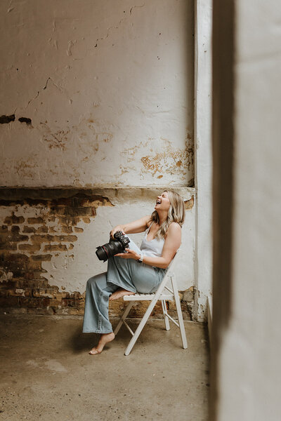 Kaz Soom, Perth Maternity, Newborn and Family Photographer.  She is sitting on a white folding chair, holding her camera and throwing her head back in laughter.