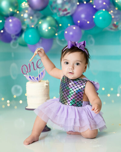 A baby girl is dressed in mermaid print and holding onto a spoon on a mermaid cake smash set