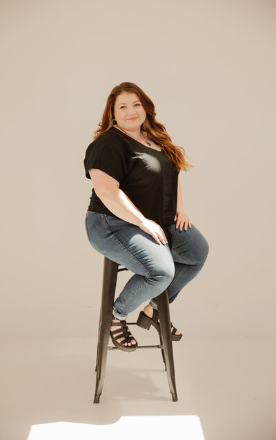 Madeline Murdock sitting posed on a stool with a white background for a brand photoshoot. Wearing a black top and jeans with curled hair and black sandals.