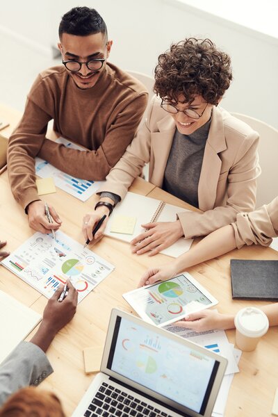 Man and woman sitting at a large table with others around going over papers with graphs on them and entering data into tablets and laptops