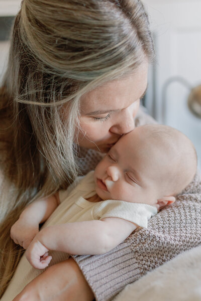 A heartfelt moment between a mother and her newborn as she gently kisses her baby’s forehead. The baby rests peacefully in her arms, wrapped in a cozy blanket, showcasing the deep bond between mother and child. Captured in soft, natural light, this image reflects the warmth and intimate storytelling of KCP Families' photography.