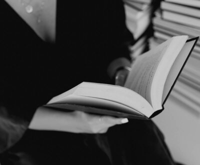 Woman in black outfit reading a book surrounded by stacked books, symbolizing knowledge and personal growth.