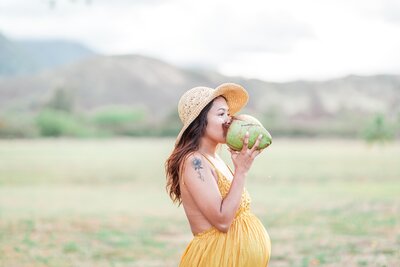 expecting mother in a mustard color dress drinking out of a coconut under wearing a sunhat in front of the koolau mountains.