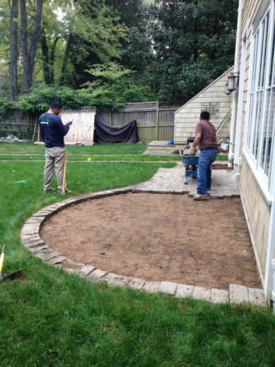 An unfinished backyard patio under construction, with workers laying a stone border and preparing the ground for paving.
