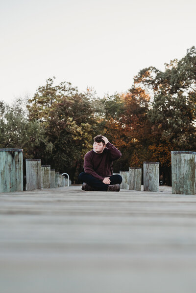 Senior photo of teen sitting on pier at sunset in red sweater and black pants.
