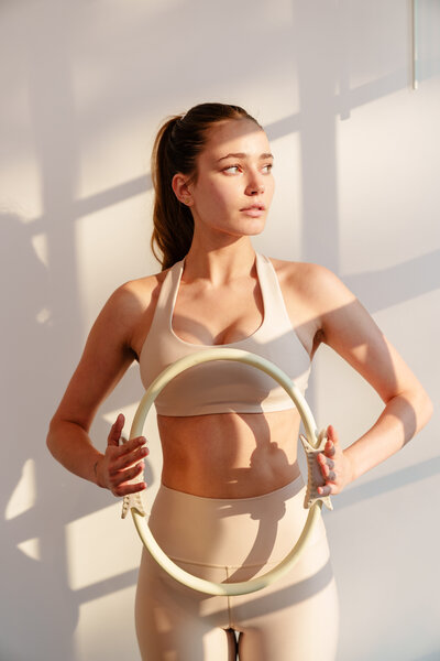 A woman in beige activewear stands in sunlight, holding a Pilates ring at waist level. She looks thoughtfully to the side, her hair in a ponytail. Shadows fall across the white wall, evoking the calm focus of a Greenwich Pilates studio.
