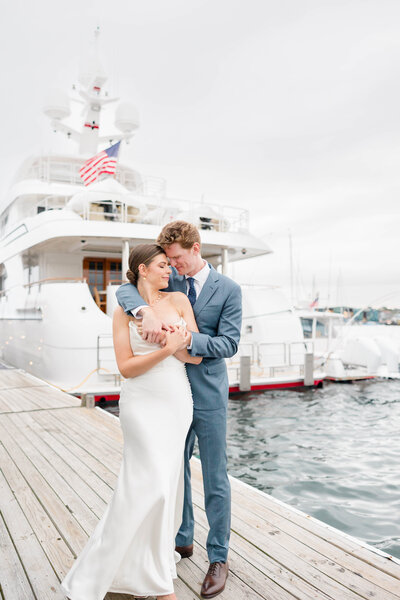 Bride and groom snuggled with each other on a deck with a white yacht behind them in Newport Rode Island CT