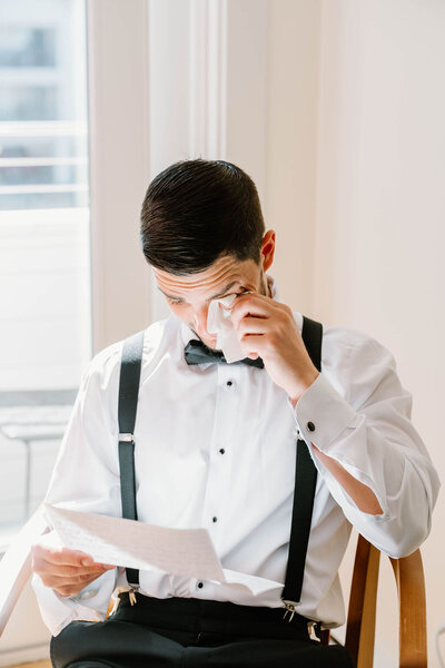Rhode Island Wedding Photographers | A man in formal attire, wearing a bow tie and suspenders, sits on a chair in soft light, wiping his tearful eyes while reading a letter with emotion.