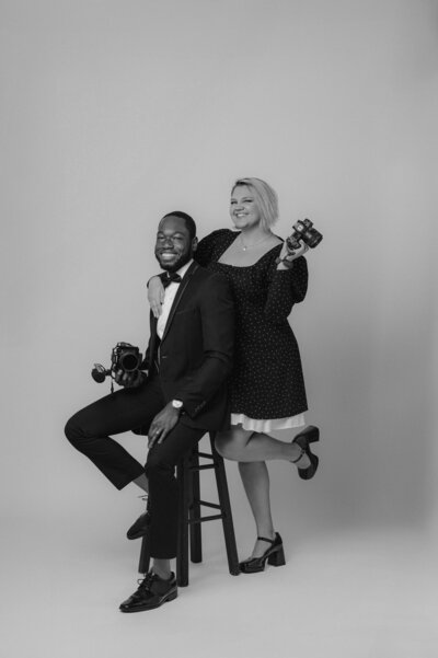 A black-and-white- studio portrait of a man and woman sitting closely together on a stool or chair against a plain backdrop.
