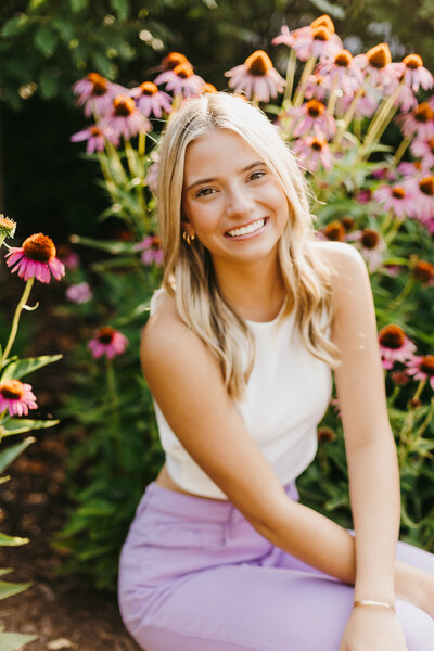 Senior girl in a white top and purple pants posing in front of pink flowers