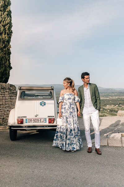 Blue and white toile off the shoulder dress and an olive green suit jacket next to a vintage citroen 2cv overlooking the valley beneath Gordes, France