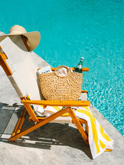 Beach bag filled with items sitting on a yellow striped towel on a chair next to the pool