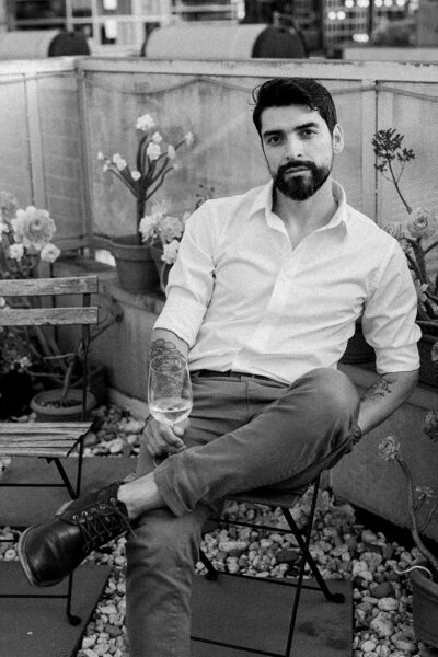 Alejandro wearing a white button down shirt, sitting on a chair holding a glass of wine