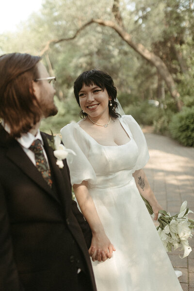 A bride and groom smile at each other while holding hands outdoors, radiating happiness.
