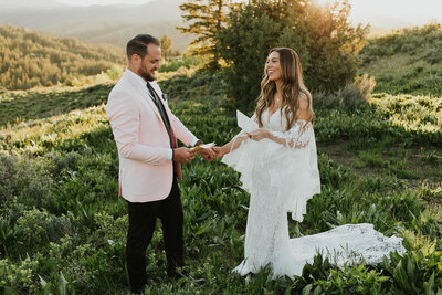 A bride and groom hold hands and read each other their vows from their books that they are holding with their free hands. They are outside on a small hill with other hills and a tree behind them. It's just them and the camera.