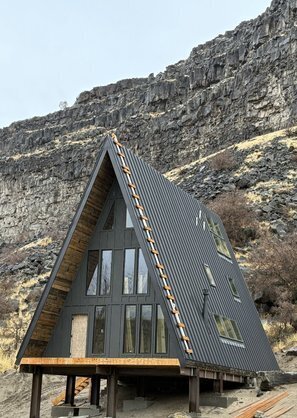 A modern A-frame cabin called the Perrine Hideaway with dark exterior siding, large glass windows, and a rugged canyon backdrop, blending nature with contemporary design.