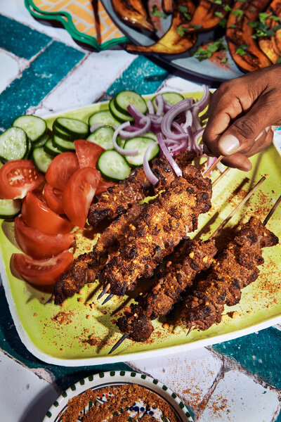 A vibrant image of grilled meat skewers served with sliced cucumbers, tomatoes, and onions on a colorful plate. Captured by Mica McCook, this photo emphasizes the rich spices and fresh ingredients, making the dish irresistible.