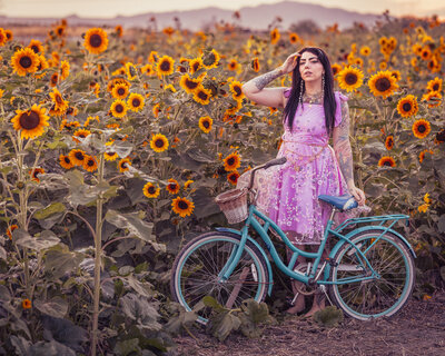 Editorial portrait photographed by Moonlit Photo in a sunflower field near Salt Lake City, featuring a woman in a lavender dress standing beside a teal vintage-style bicycle at sunset. Vibrant, artistic branding photography showcasing Utah’s natural landscapes and cinematic styling.