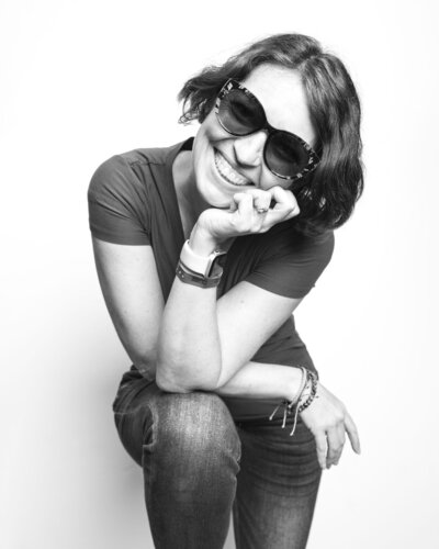 Black and white Contemorary image of woman  in sunglasses standing with one knee up and resting her elbows on the raised knee. Shot by Poplight Photography.