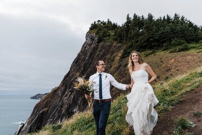 A blond bride wearing a strapless white gown holds it up while walking through the muddy trail in her hiking boots while her groom in glasses holds her waist and carries a dried floral bouquet while looking at his bride as they hike down the Elk Flats Trail near Manzanita Oregon for their adventure elopement on the coast. | Oregon Elopement Photographer