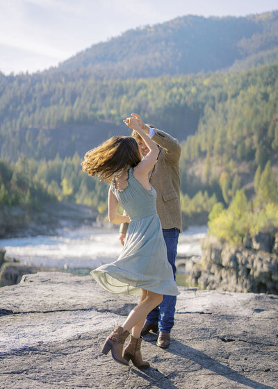 Couple dancing on a rocky cliffside with forest and river views, captured by Joseph Mayden during an outdoor adventure session in Montana.