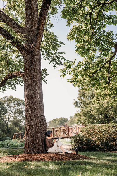 Outdoor portrait of a woman with short hair, sitting on a bench under a tree, writing in a notebook.
