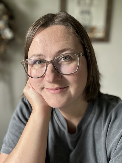 Kasey sits resting her face in her hands with a soft smile, wearing glasses and a gray shirt.