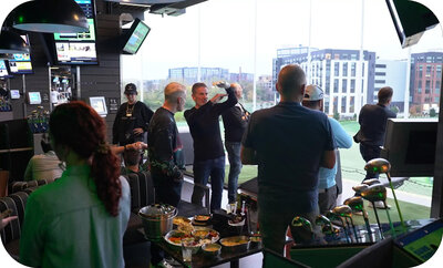 Group of people gathered at an indoor golf venue, talking and watching someone take a swing near the hitting bay.