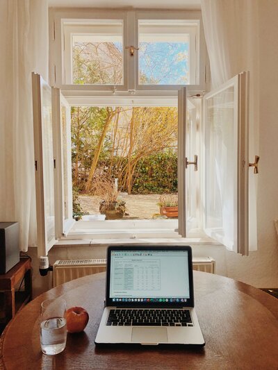 A laptop on a wooden desk, looking out an open window with a sunny view.